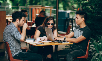 This image is of people sitting outside a restaurant with branches hanging over and 3 people are sitting at a table in front of the image.